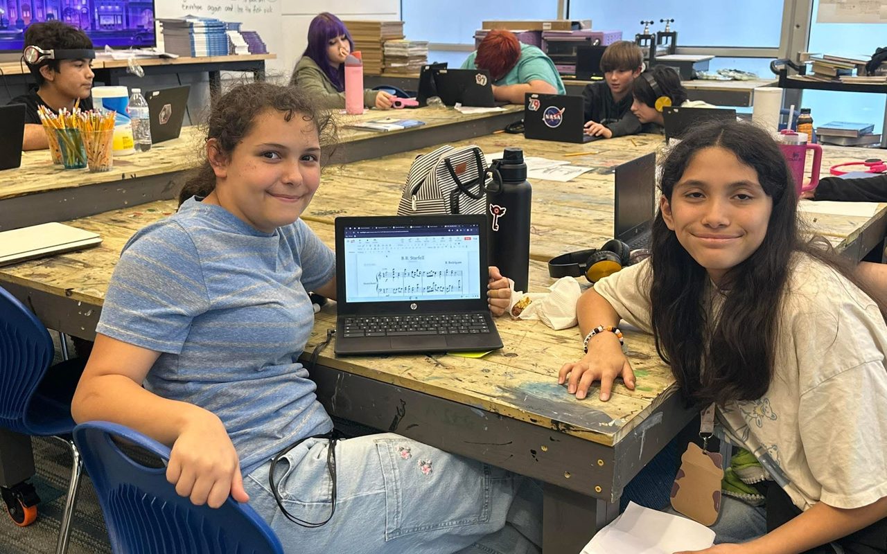 Two TriPoint students smiling together at a classroom table with a laptop.